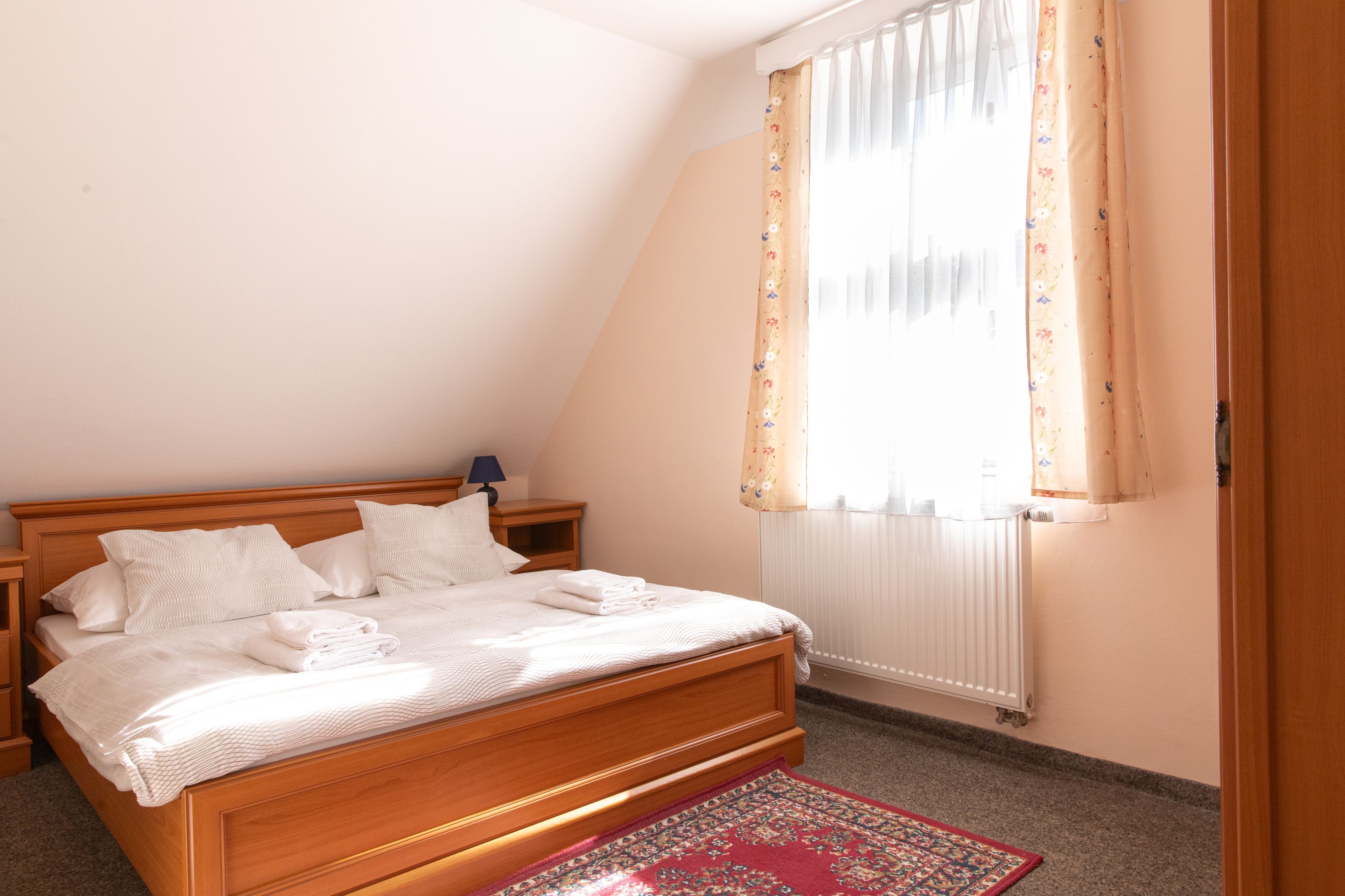 A bedroom with a wooden bed, white bedding, a nightstand with a lamp, a window with floral curtains, and a radiator.