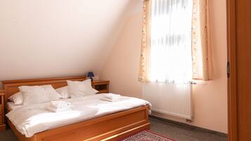 A bedroom with a wooden bed, white bedding, a nightstand with a lamp, a window with floral curtains, and a radiator.