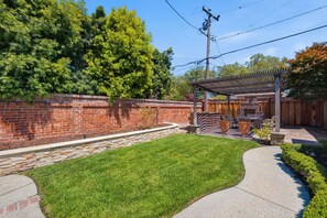 A backyard with a brick wall, a grassy lawn, a pergola, and a wooden deck. - Modern Fully Furnished House - 3 Bed 2 Bath King Bed Pool & Jacuzzi (San Jose)