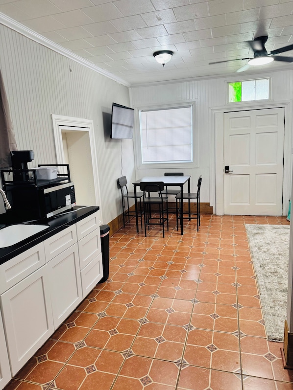 A kitchen with white cabinets, a black countertop, a tiled floor, a ceiling fan, a window with blinds, and a door.