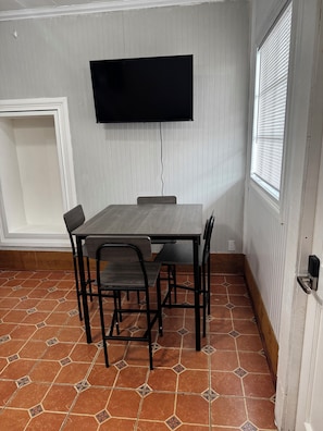 A room with a tiled floor, a table with chairs, a mounted television, and a window with blinds. - Charming 1-bedroom cottage in Ruston located perfectly for your convenience (Ruston)