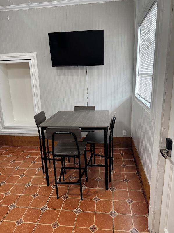 Una habitación con piso de baldosas, una mesa con sillas, televisor montado en la pared y una ventana con persianas.