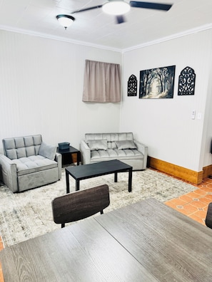 A living room with a grey sofa, a smaller grey armchair, a black coffee table, and a wooden dining table. There are two wall-mounted artworks depicting a forest scene and a decorative metal piece. - Charming 1-bedroom cottage in Ruston located perfectly for your convenience (Ruston)