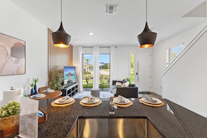 A modern living room with a kitchen area, a dining table set for four, and a large window offering a view of the outdoors. - 3BR Near Disney, Harry Potter and Mickey Rooms (Kissimmee)