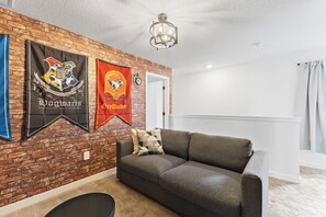 A room with a brick wall, a ceiling fan, a grey sofa, and two banners. - 3BR Near Disney, Harry Potter and Mickey Rooms (Kissimmee)