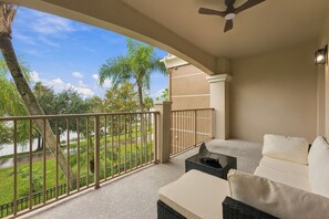 A balcony with a view of a grassy area and trees, a ceiling fan, and a sofa with cushions. - Luxury 3br, Modern Design, Lake Views, Vista Cay (Orlando)