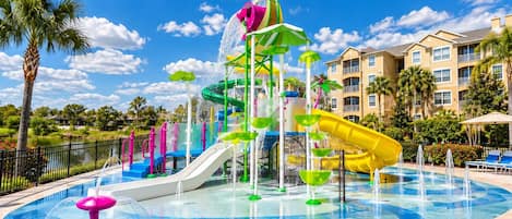 A water park with slides and fountains, surrounded by a pool and palm trees.