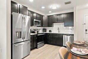 A modern kitchen with stainless steel appliances, dark cabinetry, and a dining area with a table set for two. - Spacious 3br, Top Location Near Parks & Convention (Orlando)