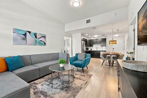 A modern living room with a grey sofa, blue armchair, and a coffee table with a plant. A kitchen is visible in the background with dark cabinets and stainless steel appliances. - Spacious 3BR Condo, Perfect Orlando Home Base (Orlando)