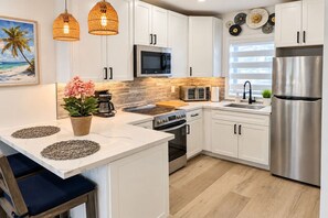 A modern kitchen with white cabinets, a stainless steel refrigerator, a microwave, and a stove. There is a potted plant on the counter and a painting of a beach on the wall. - Steps to Hollywood Beach - Modern 1BR Retreat (Hollywood)