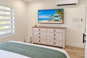 Une chambre à coucher comprenant un lit, une commode, un téléviseur fixé au mur et une fenêtre avec des stores.