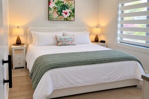 Une chambre à coucher avec un lit, deux tables de chevet, une horloge murale, une fenêtre avec des stores et un tableau représentant des fleurs.