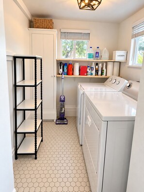 A laundry room with a washing machine, a vacuum cleaner, shelves with cleaning supplies, and a basket on the wall. - Historic Home: Walk to Downtown, Jays & Waterfront (Dunedin)
