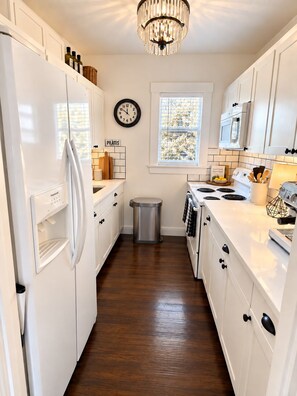 A kitchen with white cabinets, a stainless steel trash can, a clock, and a window with patterned blinds. - Historic Home: Walk to Downtown, Jays & Waterfront (Dunedin)