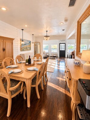 A dining area with a wooden table set for four, chairs, a chandelier, and a view of the outdoors through large windows. - Historic Home: Walk to Downtown, Jays & Waterfront (Dunedin)