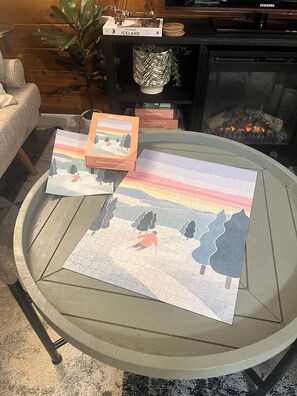 Une table basse ronde en verre, sur laquelle se trouvent un jeu de puzzle, un livre intitulé « L’Islande, terre de contrastes », et un objet décoratif posé sur une surface en bois.