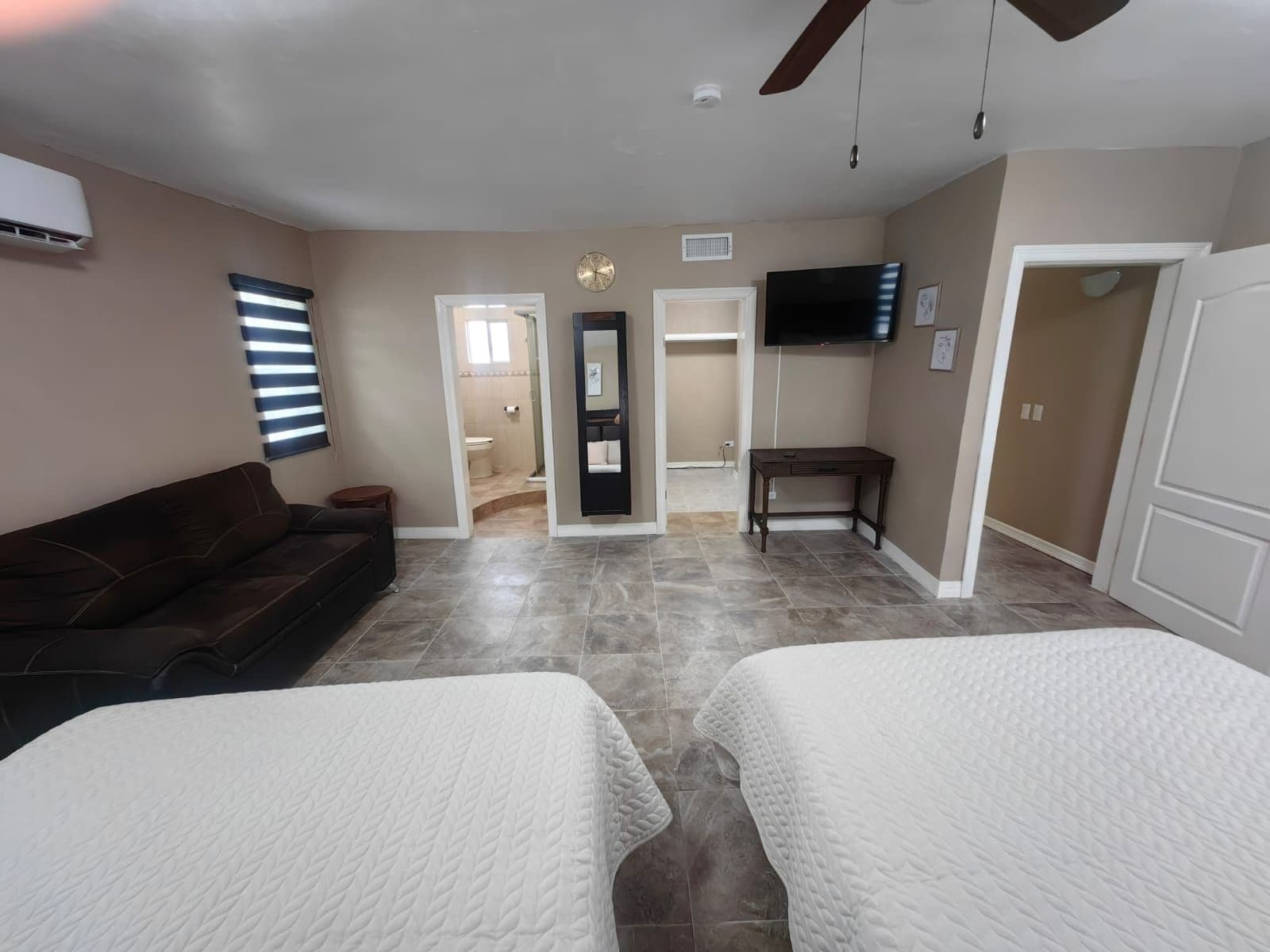 A hotel room with a brown sofa, two beds with white bedding, a flat-screen TV, a ceiling fan, and a bathroom visible through an open door.