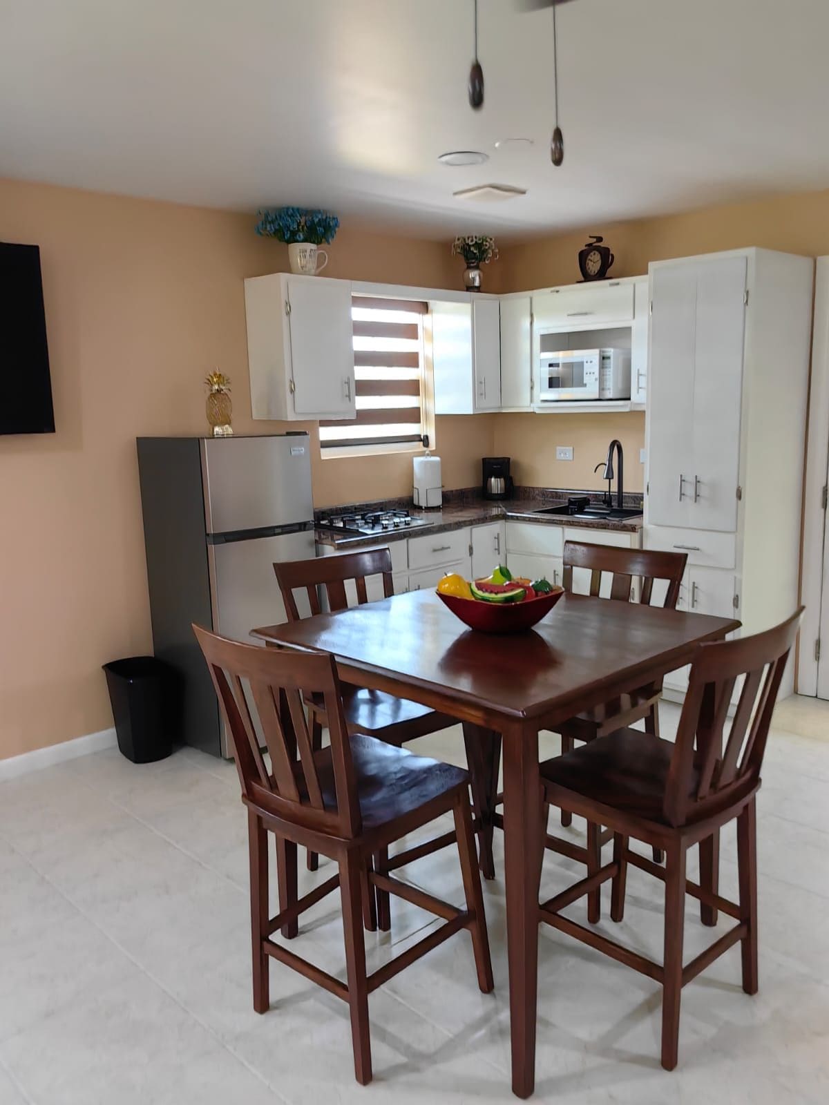 A dining area with a table, chairs, a bowl of fruit, and a kitchen in the background.