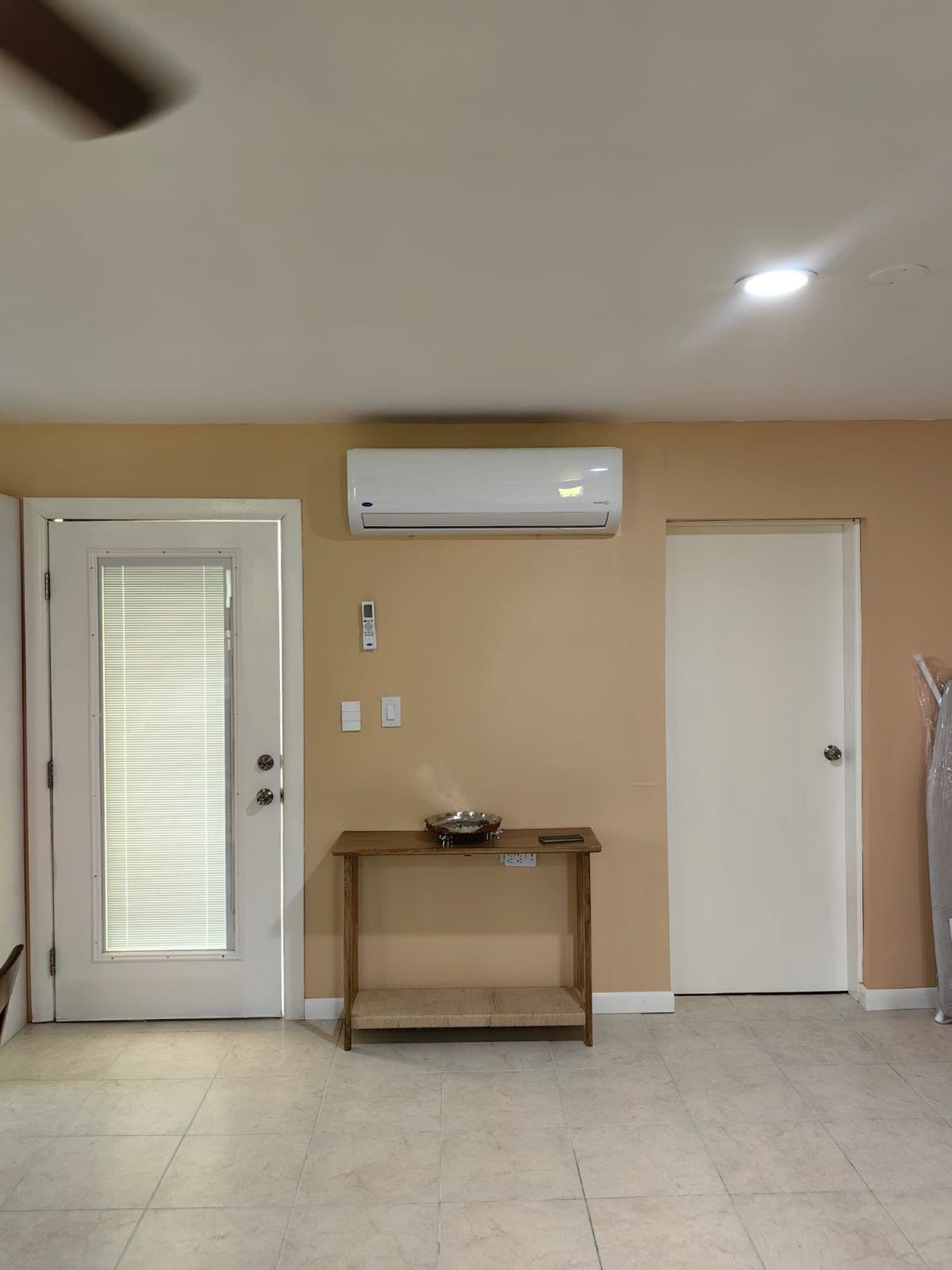 Una habitación con paredes color beige, una puerta blanca con persianas, un ventilador de techo blanco y un equipo de aire acondicionado blanco montado en la pared.