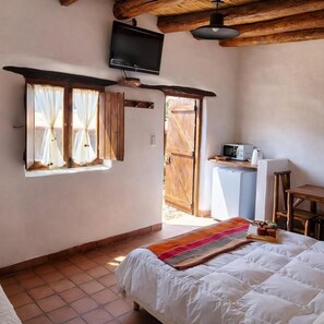 Un dormitorio con cama, televisor montado en la pared, ventana con cortinas y una pequeña zona de kitchenette.