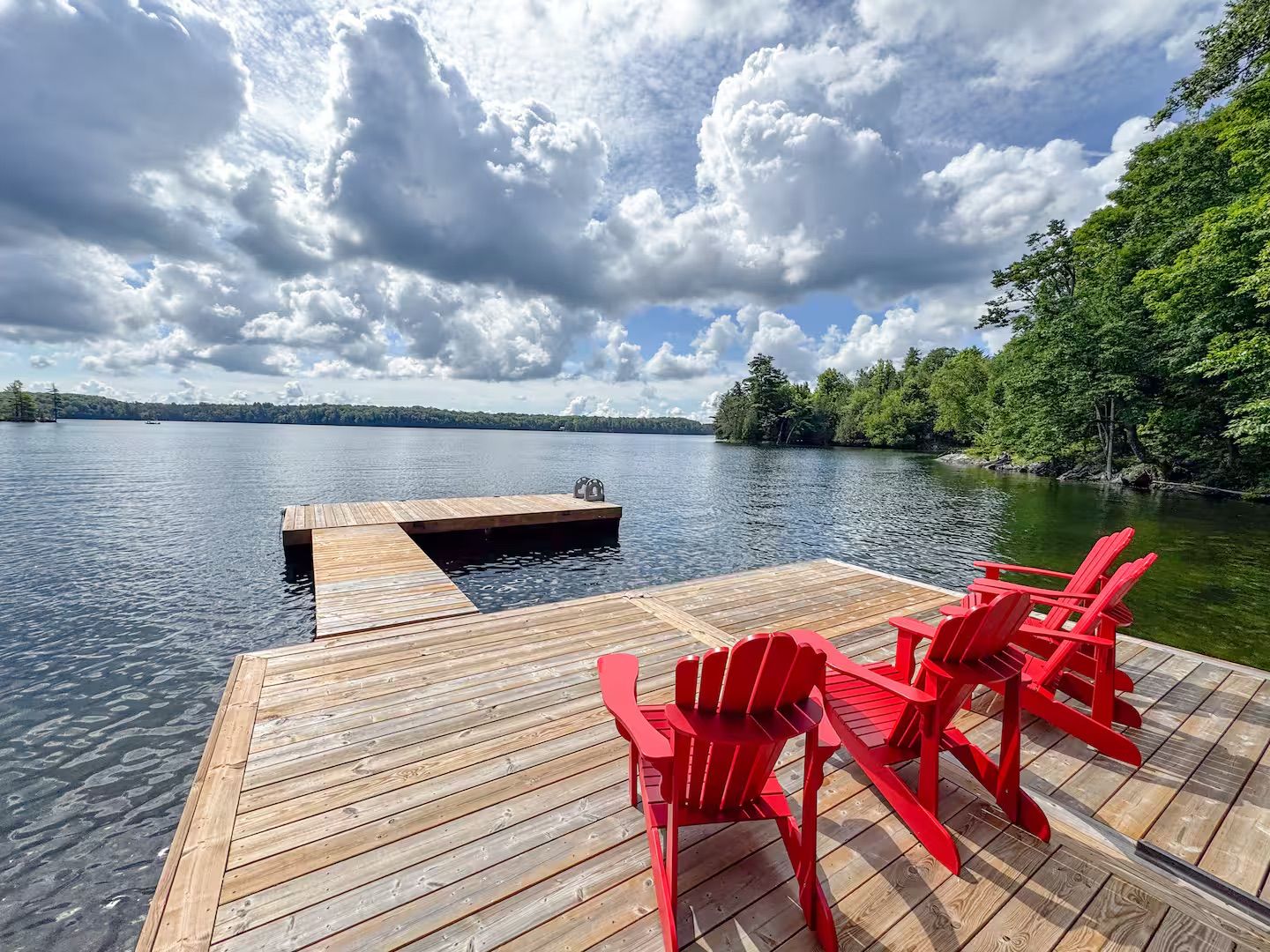 A wooden dock with a table and red chairs extending over a lake, surrounded by trees.