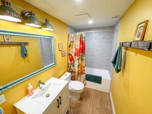 Un bagno con lavandino, WC e vasca da bagno bianchi. Si vede uno specchio con cornice decorativa, una tenda da doccia con motivo floreale e un portasciugamani con un asciugamano verde.
