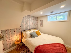A bedroom with a bed, a window, a wall with a birch tree pattern, and a small table with a lamp.