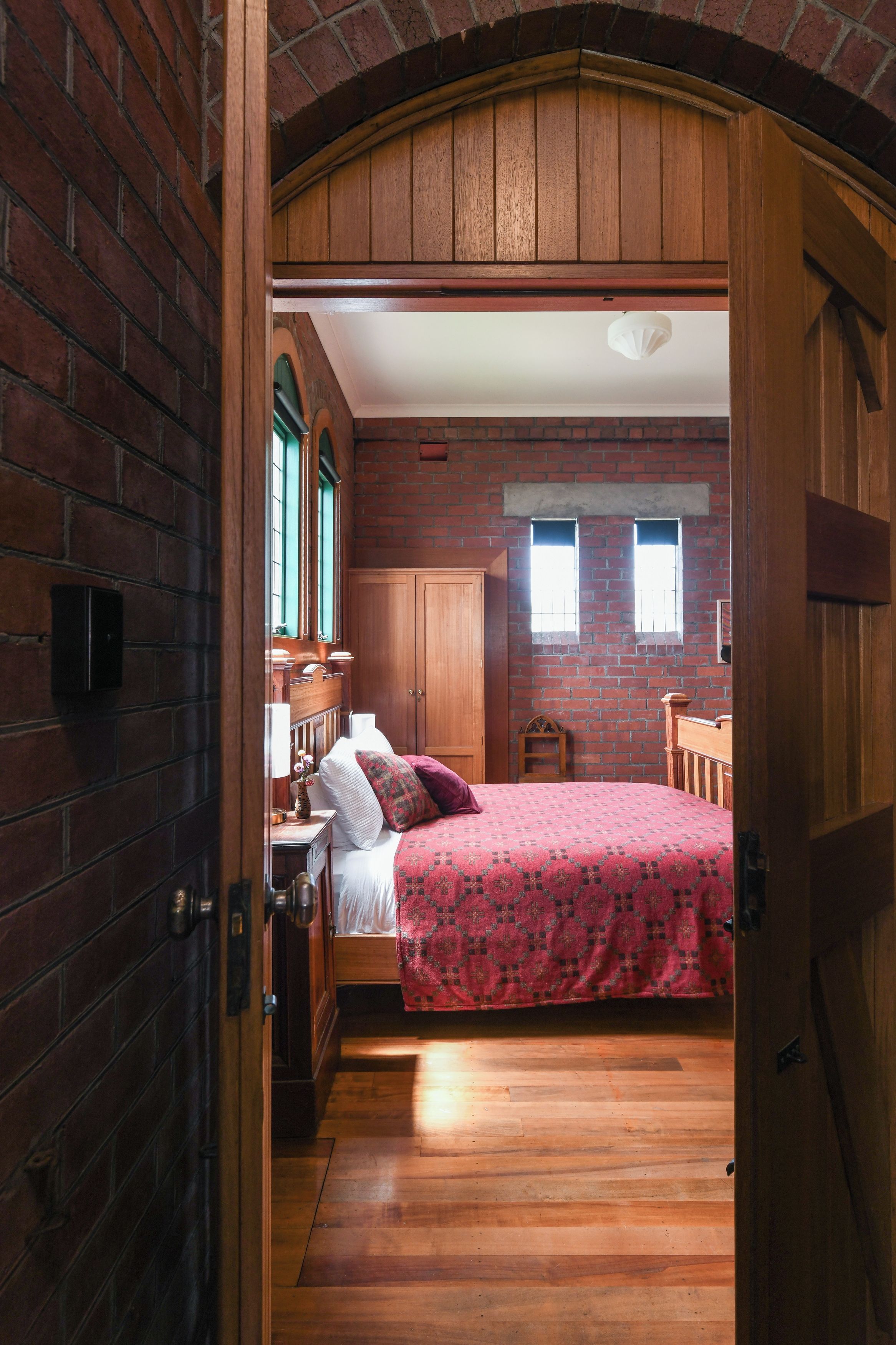 Un dormitorio con puerta de madera, una cama con colcha estampada, una mesita de noche, una lámpara y una ventana con vista a un muro de ladrillo.