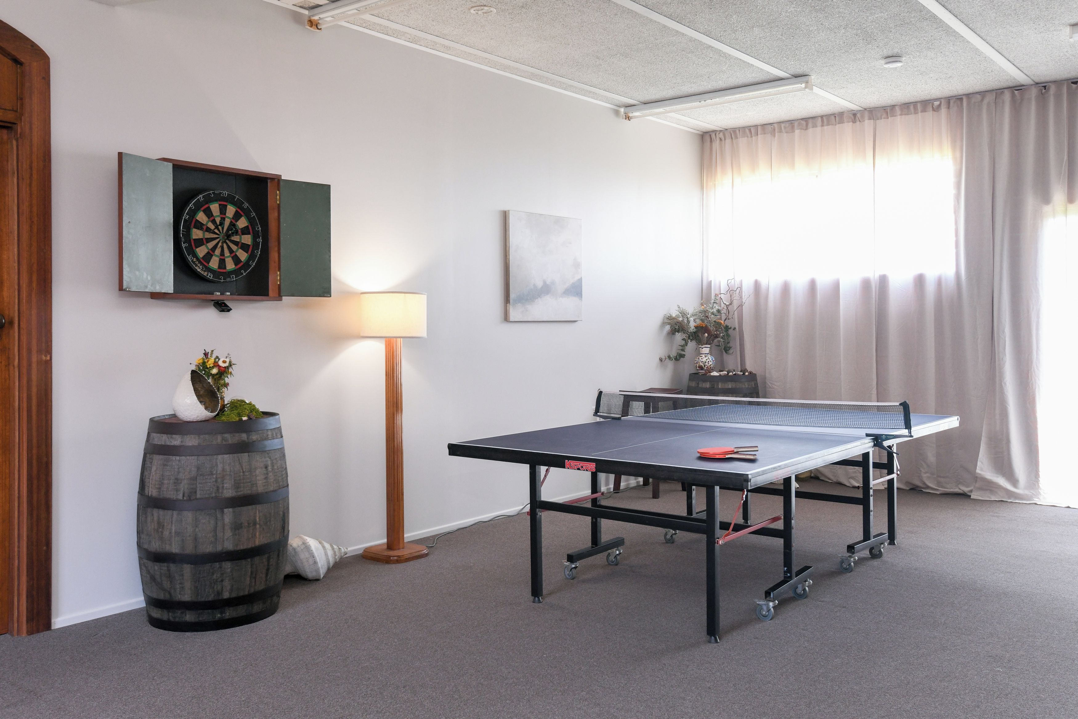 Una habitación con mesa de ping pong, dardo, lámpara y un amplio ventanal con cortinas.