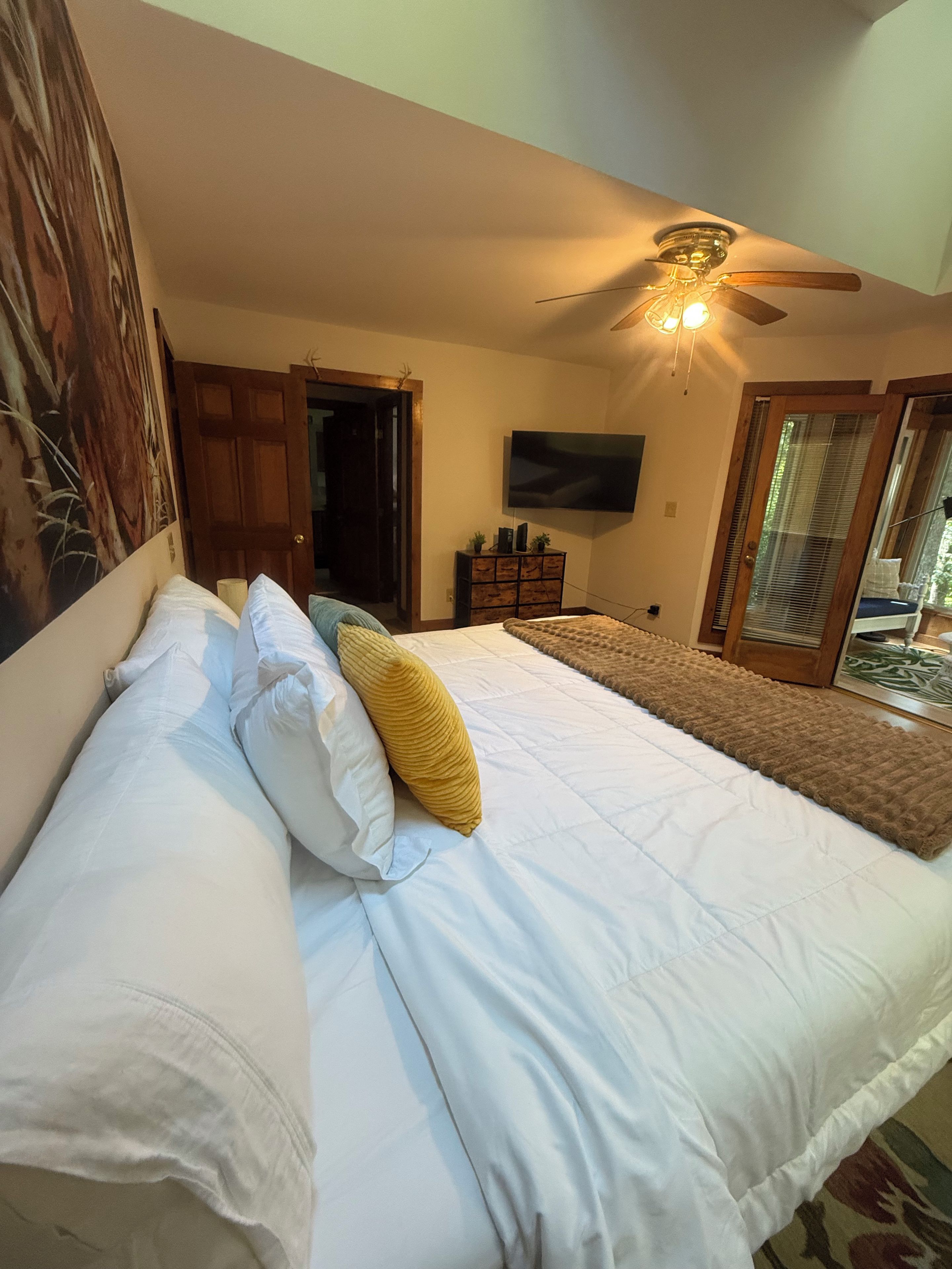 A bedroom with a bed, a ceiling fan, a television, and a door leading to a balcony.
