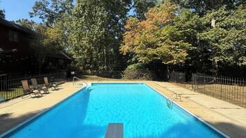 A rectangular swimming pool with a concrete deck in the foreground, surrounded by a metal fence and trees with green and autumnal foliage.