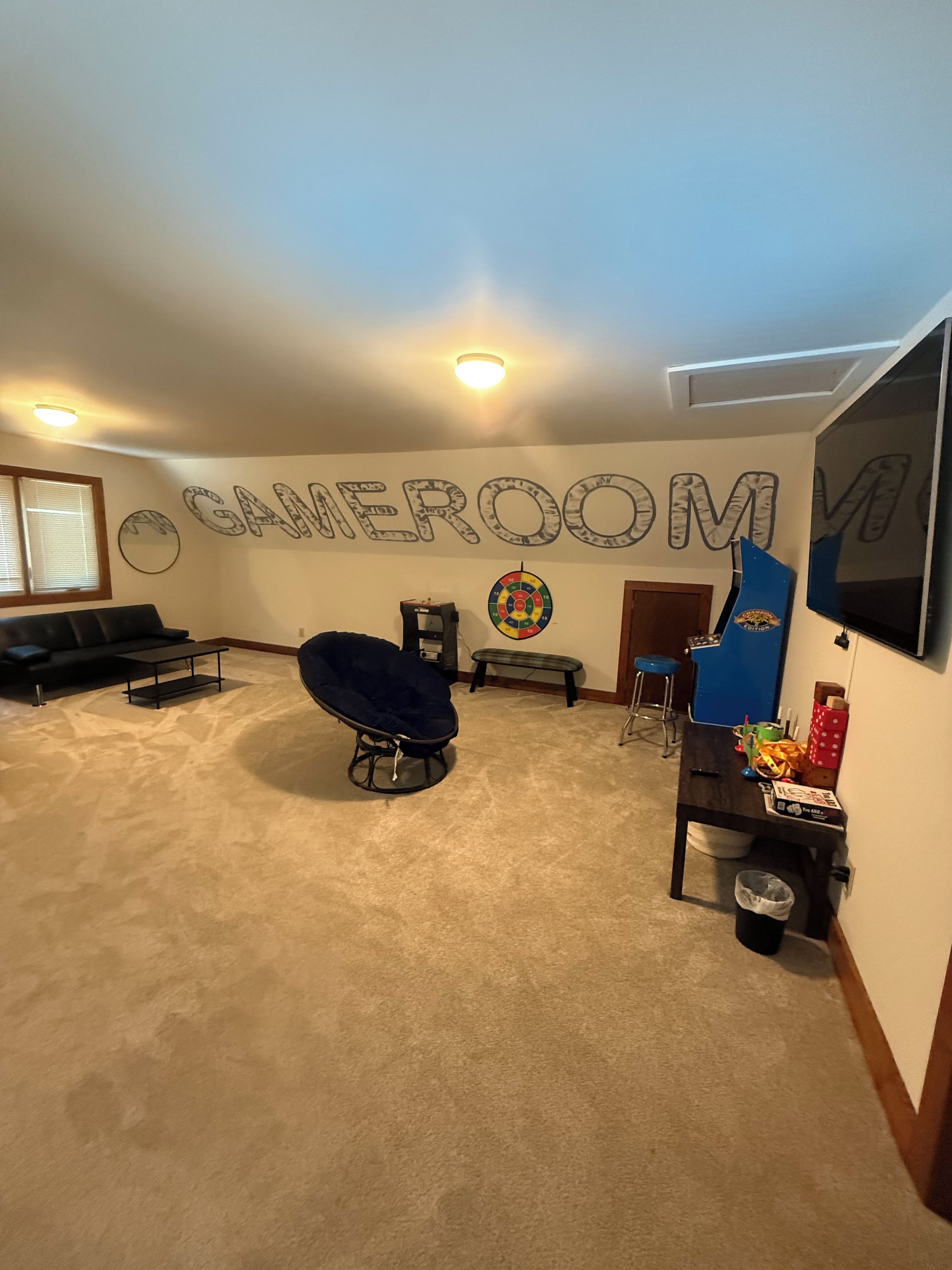 Una habitación con un gran letrero en la pared que dice 'GAMEROOM', un sillón de peluche, un dardo, una mesita y un televisor montado en la pared.