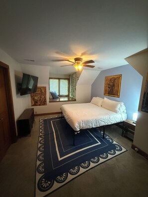 Un dormitorio con una cama, ventilador de techo, televisor, ventana con persianas y un cuadro en la pared.