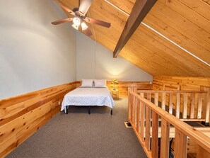 Un dormitorio con techo de madera, una cama con edredón blanco, una mesita de noche con lámpara y una barandilla de madera.