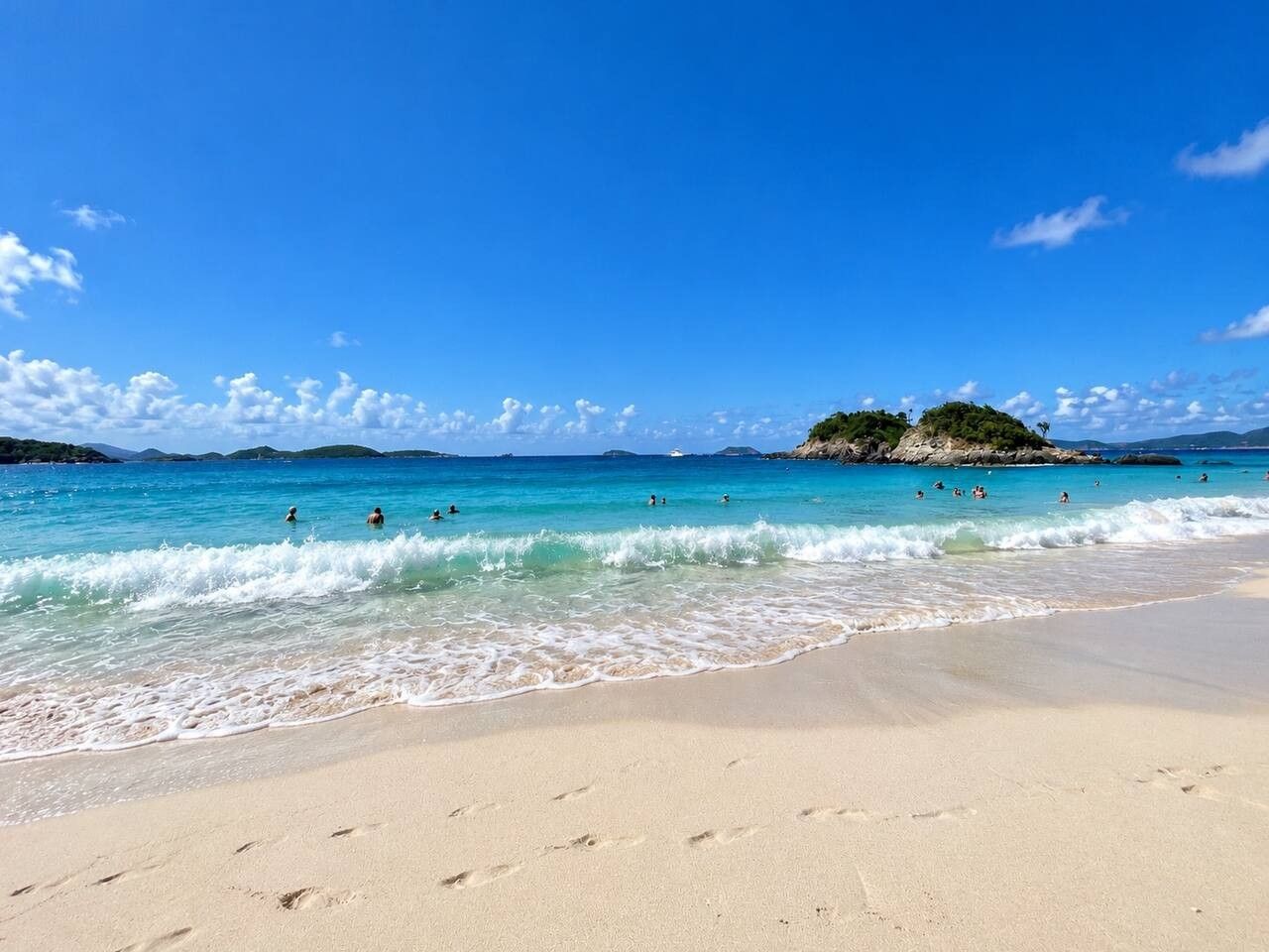 Une plage avec une eau d’un bleu limpide, des gens qui se baignent et une rive sablonneuse.