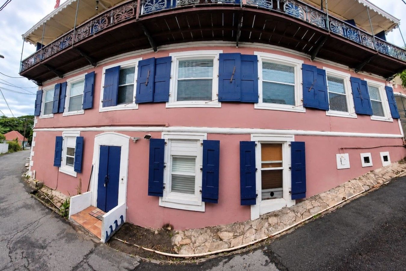 Ein rosafarbenes Gebäude mit blauen Fensterläden, einem Balkon mit aufwendiger Railing und einem Steinfundament.
