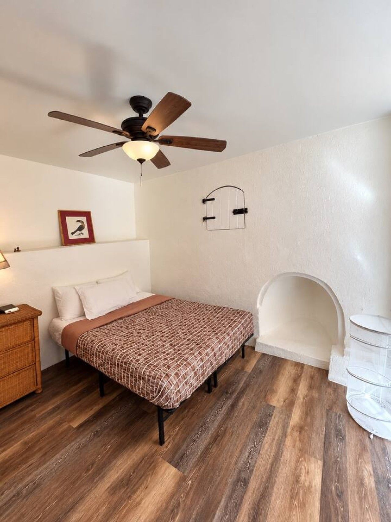 A bedroom with a bed, a wooden floor, a ceiling fan, a framed picture, a wall-mounted light, and a white arched window.
