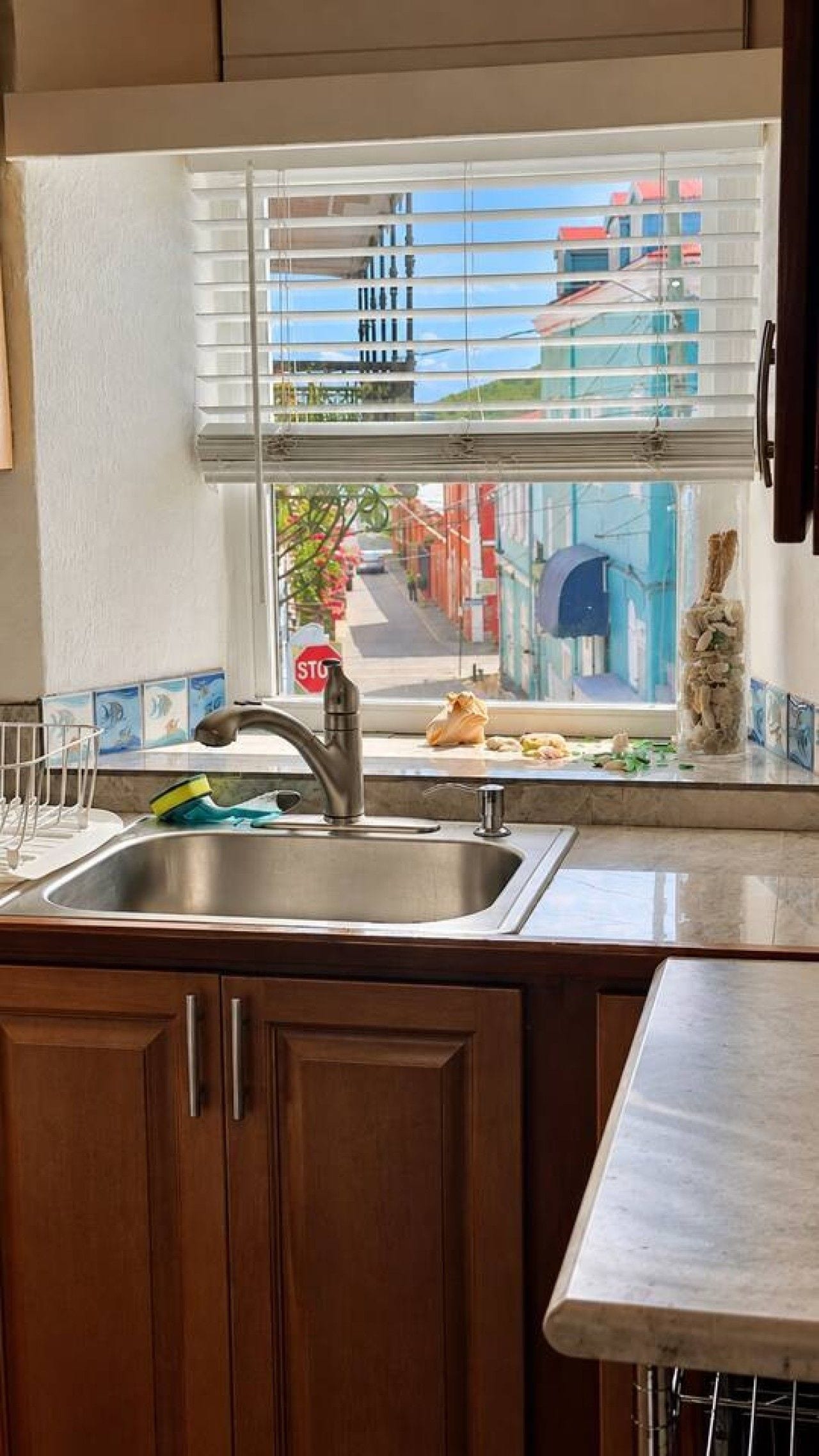 A kitchen with a stainless steel sink, wooden cabinets, a window with blinds, and a view of buildings outside.