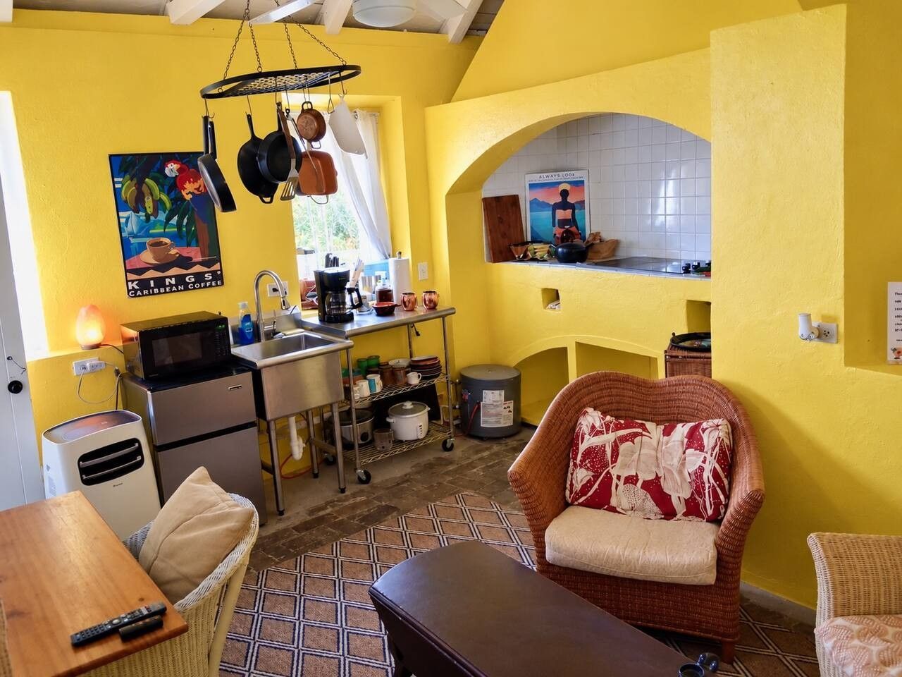 A compact kitchen with a microwave, stove, and hanging pots. There is a wicker chair with a cushion and a small table with a remote control.