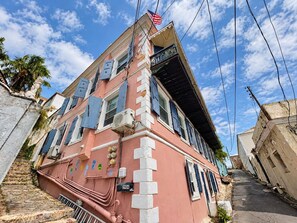 A multi-story building with a pink facade, a visible American flag, and a narrow street leading to it. - King’s Lighthouse | Garden Retreat, Downtown (Charlotte Amalie)