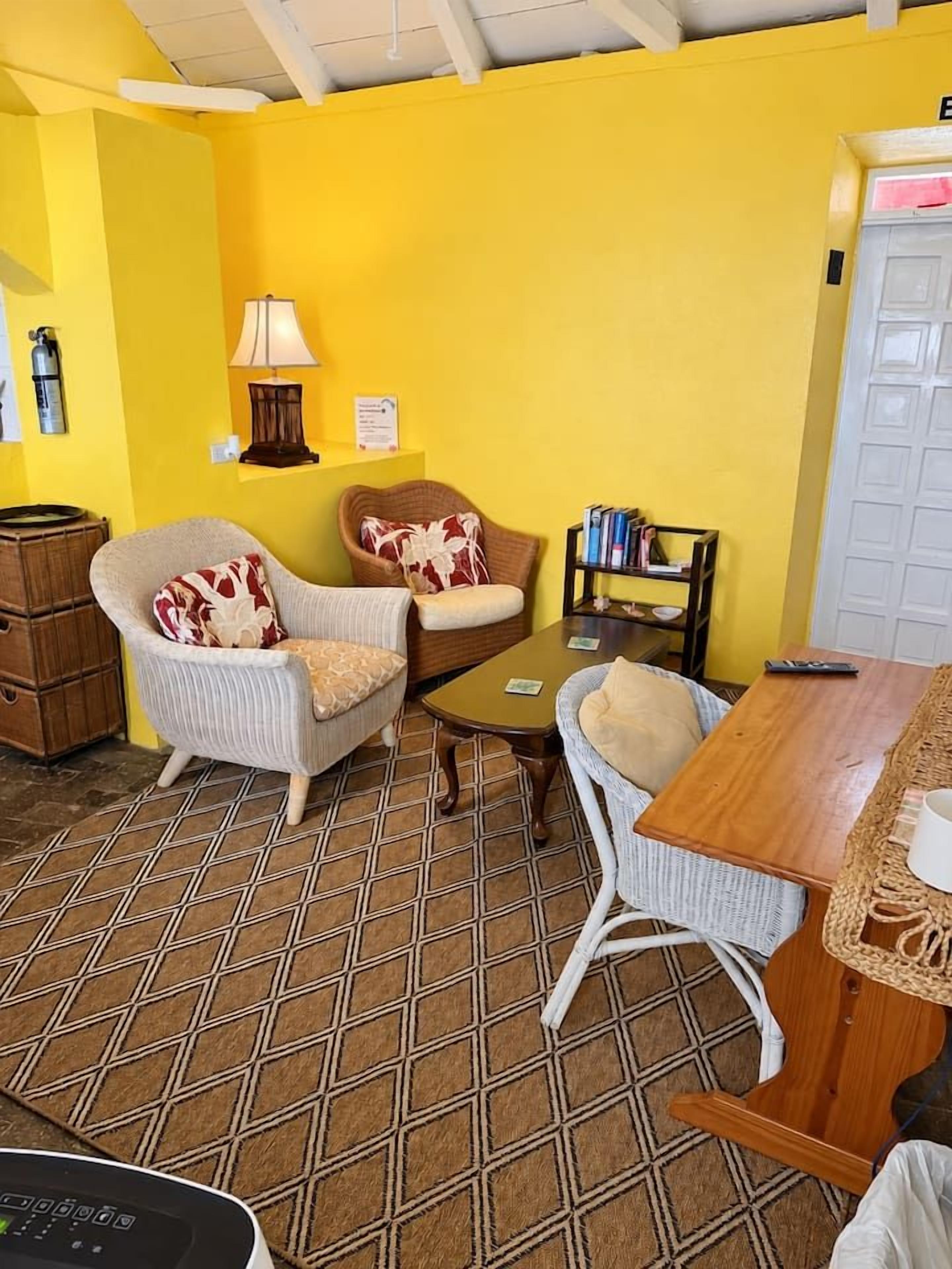 Une pièce avec un mur jaune, une table en bois, deux chaises en osier, une petite bibliothèque et un extincteur.