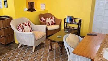 Une pièce avec un mur jaune, une table en bois, deux chaises en osier, une petite bibliothèque et un extincteur.