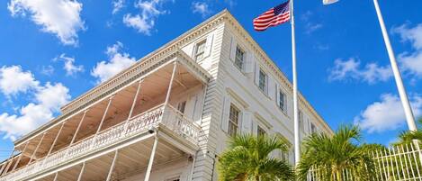 A white building with a balcony, an American flag, and a flag with a blue field and a gold eagle.