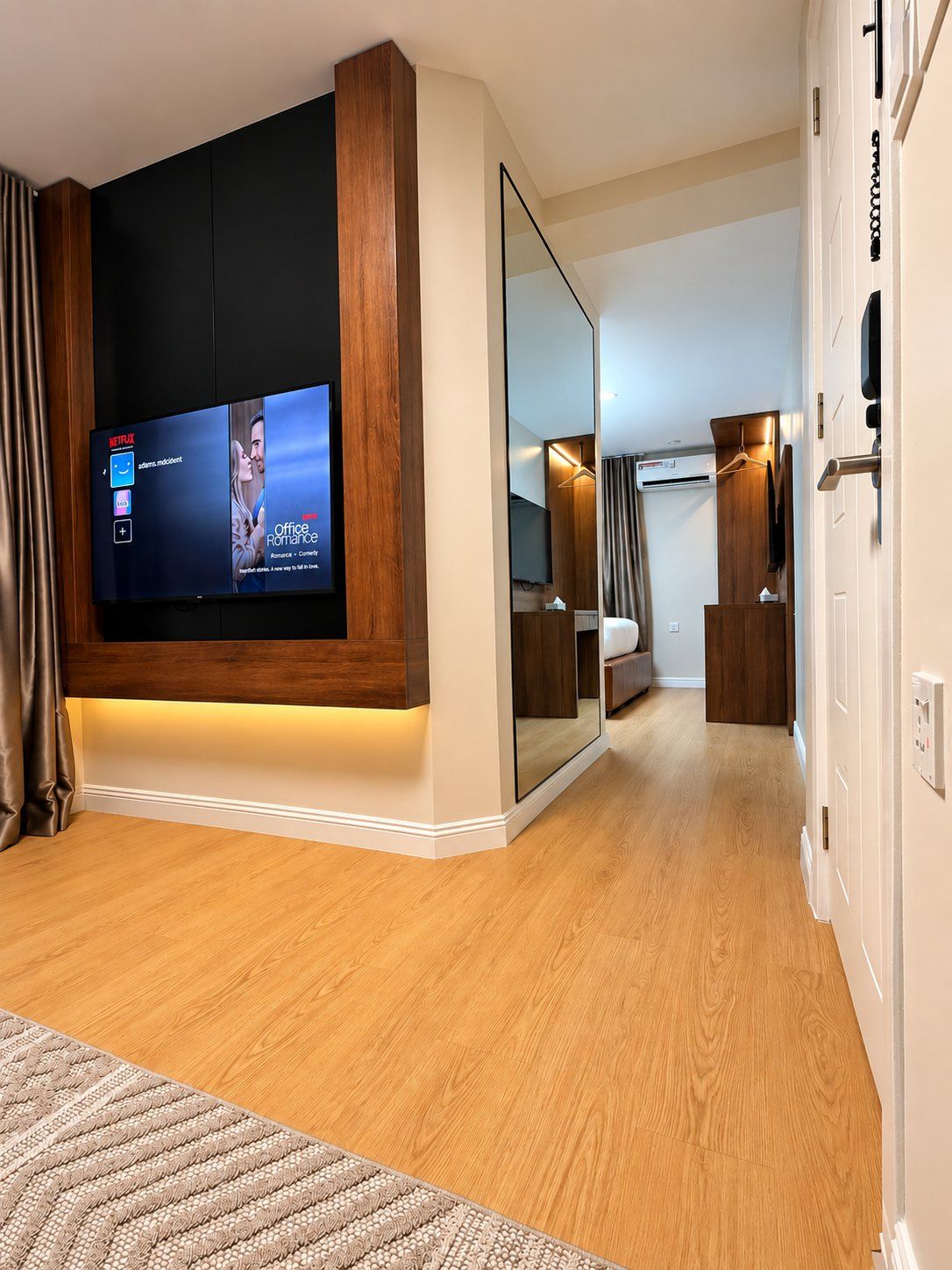 Une chambre d’hôtel moderne dotée d’un grand téléviseur à écran plat fixé au mur, d’un dressing et d’un couloir menant à une autre chambre.