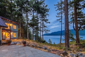 A two-story house with a stone patio, surrounded by tall trees and overlooking a calm lake. - Private Luxury 4 Bedroom Estate on 4 Acres with over 1,100 feet of shoreline! (Orcas)