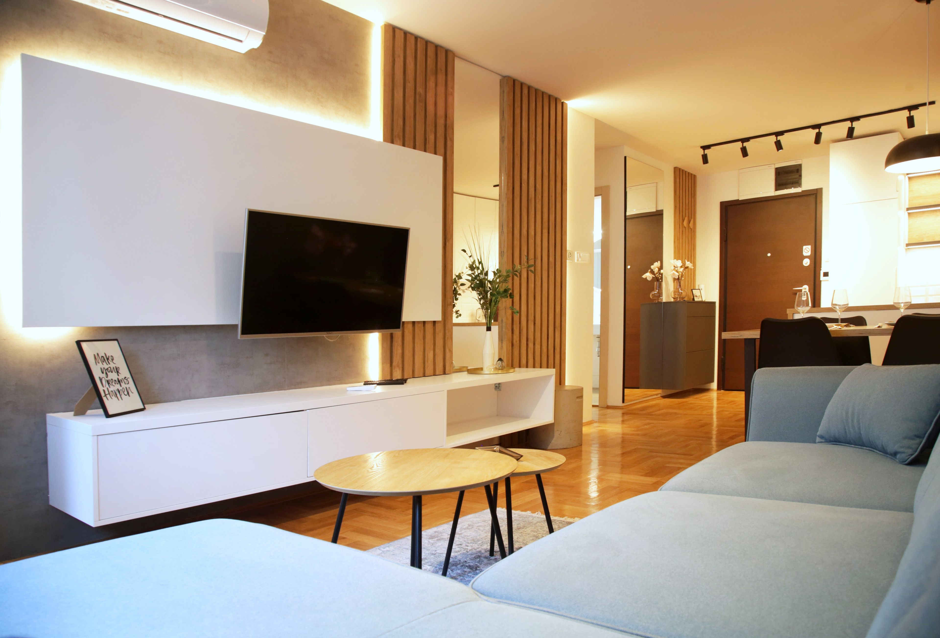 A modern living room with a flat-screen TV mounted on a white console, a round wooden coffee table, and a light grey sofa.