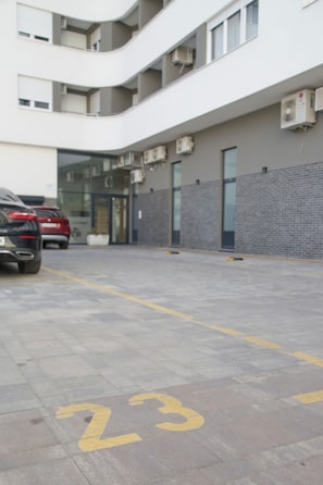 Ein Parkplatz mit markierten Stellplätzen, ein Gebäudeeingang und Klimaanlagen an der Außenwand.