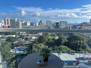 A balcony with a view of a city skyline, a potted plant, and a building with signage. - Apartment with an ocean view in the heart of the city (Santo Domingo)