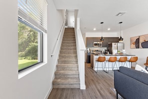 Een modern interieur met een trap die leidt naar een keuken, een eethoek met barkrukken, en een zithoek met een bank.