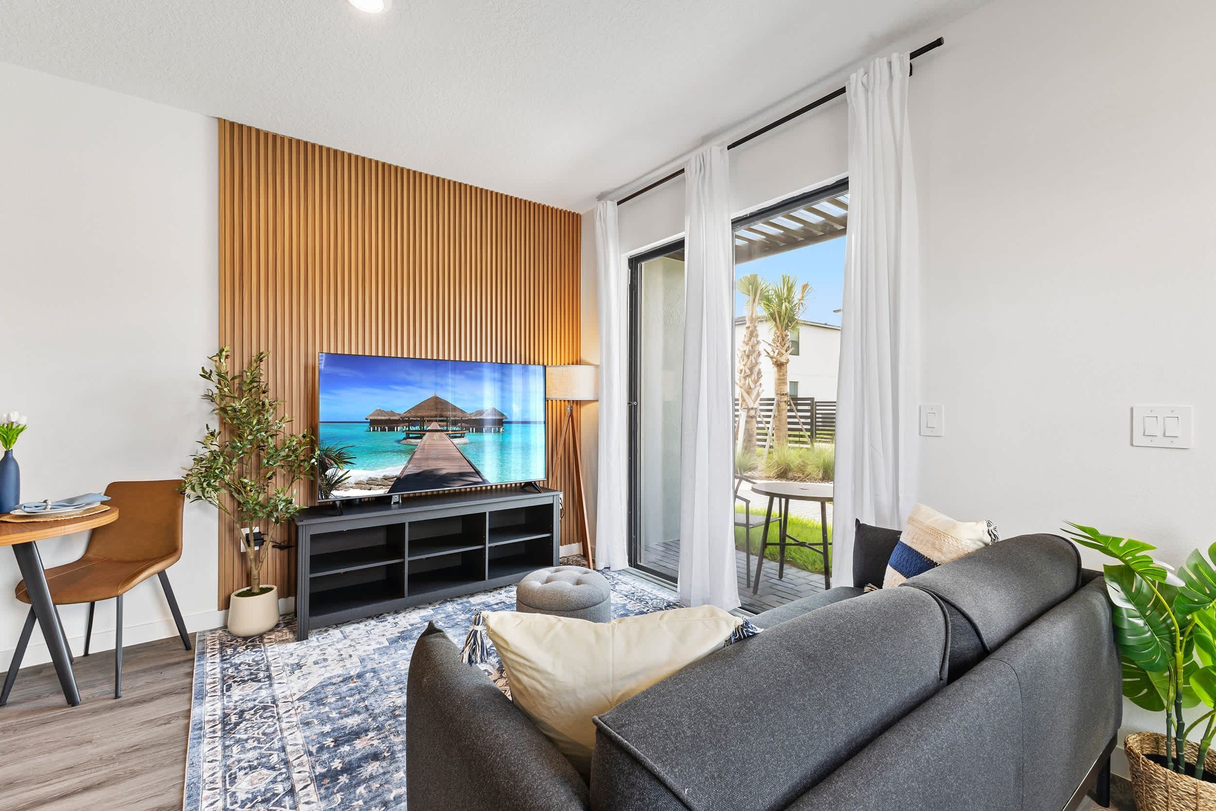 A modern living room with a flat-screen TV, wooden panel wall, and a view of a tropical beach through the glass door.